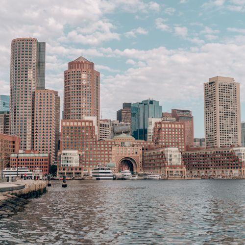 The image shows a city skyline with tall buildings and a waterfront, with boats docked by the pier, under a partly cloudy sky.