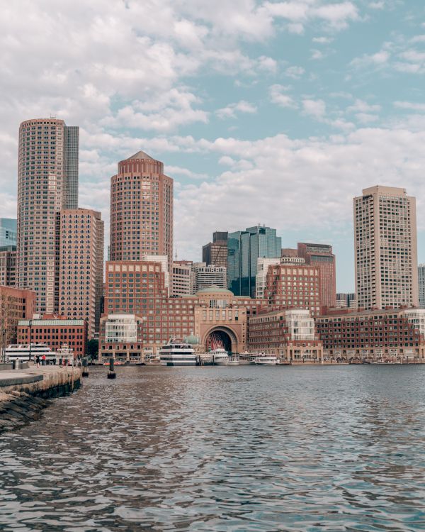 The image shows a city skyline with tall buildings and a waterfront, with boats docked by the pier, under a partly cloudy sky.