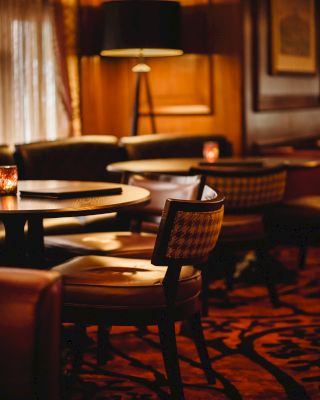 The image shows a cozy, dimly lit café or restaurant with wooden furniture, cushioned chairs, a patterned carpet, and a floor lamp in the background.