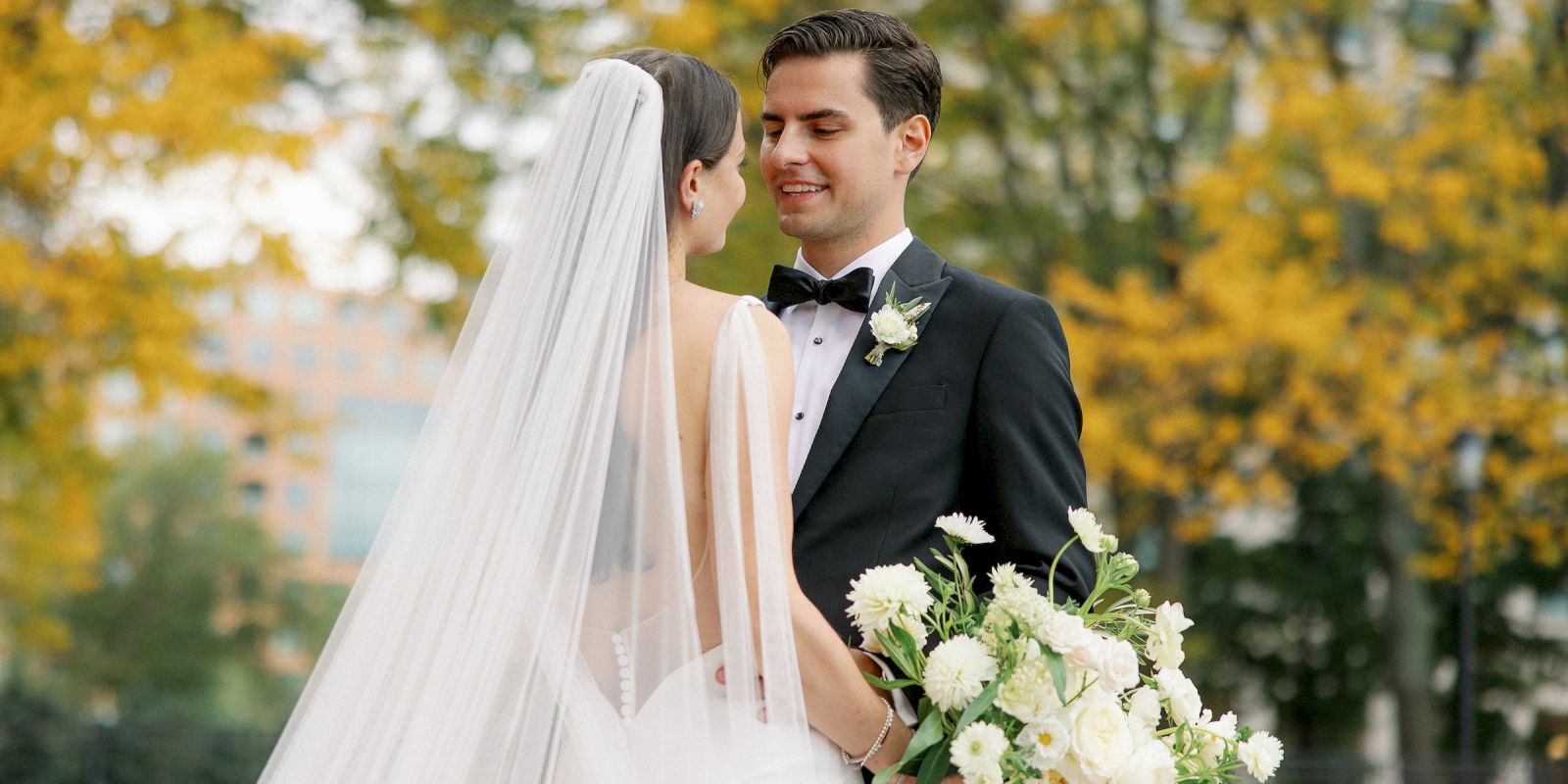 A bride and groom share a moment outdoors, surrounded by autumn trees; the bride holds a bouquet, and both appear joyful.