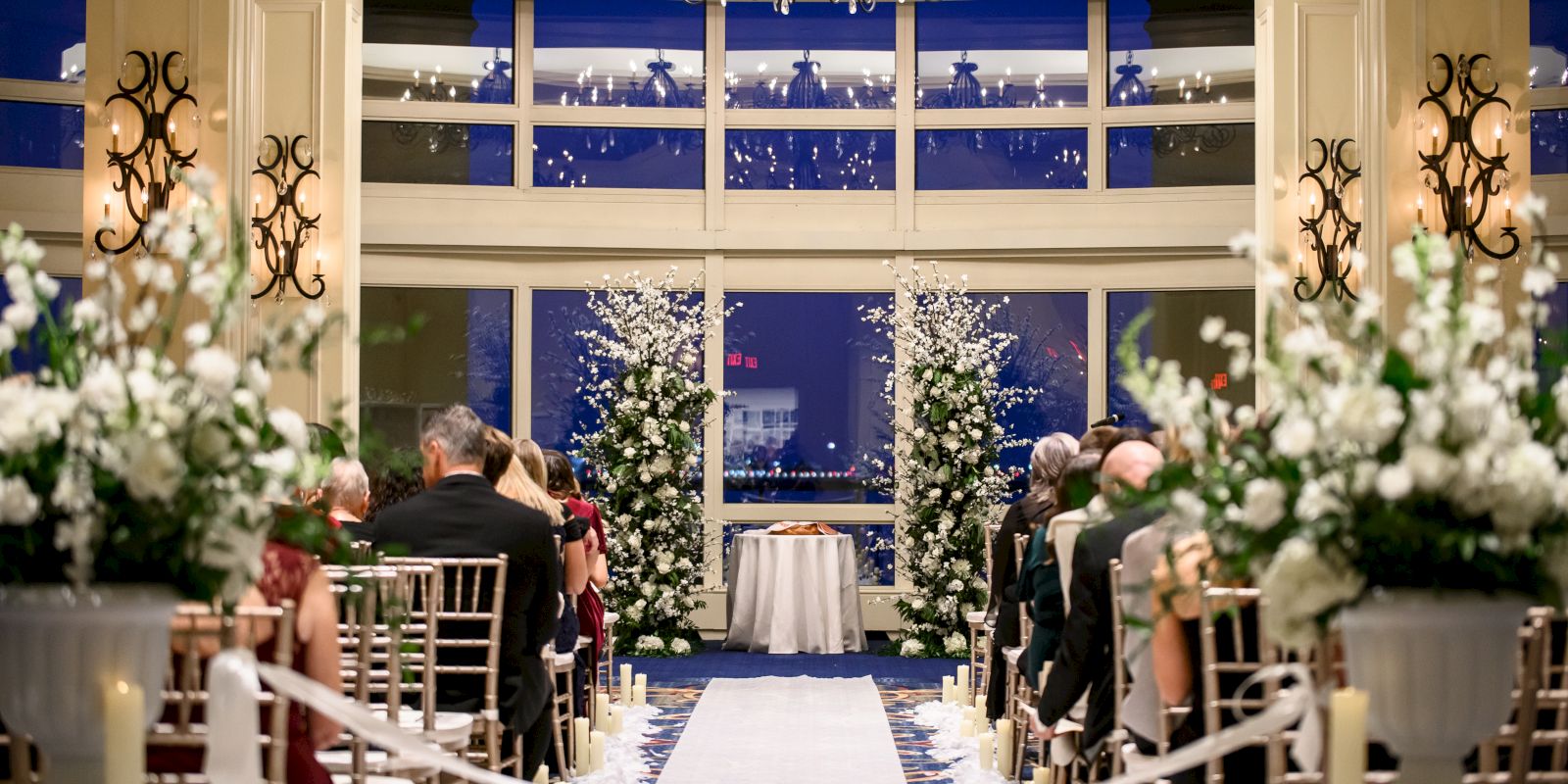 An indoor wedding ceremony setup with chairs, floral arrangements, and an aisle leading to an altar, with large windows in the background.