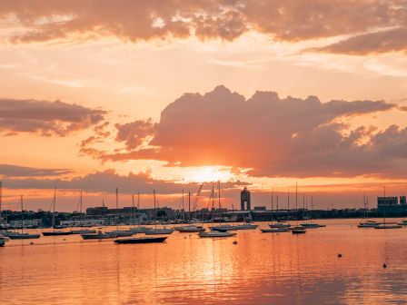 Sunset over water with clouds, boats, and a distant lighthouse.