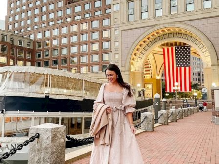 A woman stands in front of an urban backdrop with buildings and a large American flag.