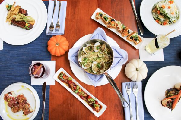 A table is set with various dishes, drinks, and small decorative pumpkins on a wooden surface, featuring pasta, bruschetta, and salad.