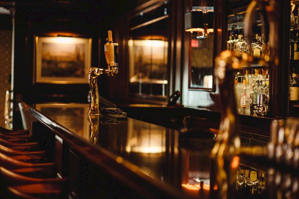 This image shows an empty bar counter with a golden beer tap, wooden bar stools, and shelves stocked with various bottles of alcohol.