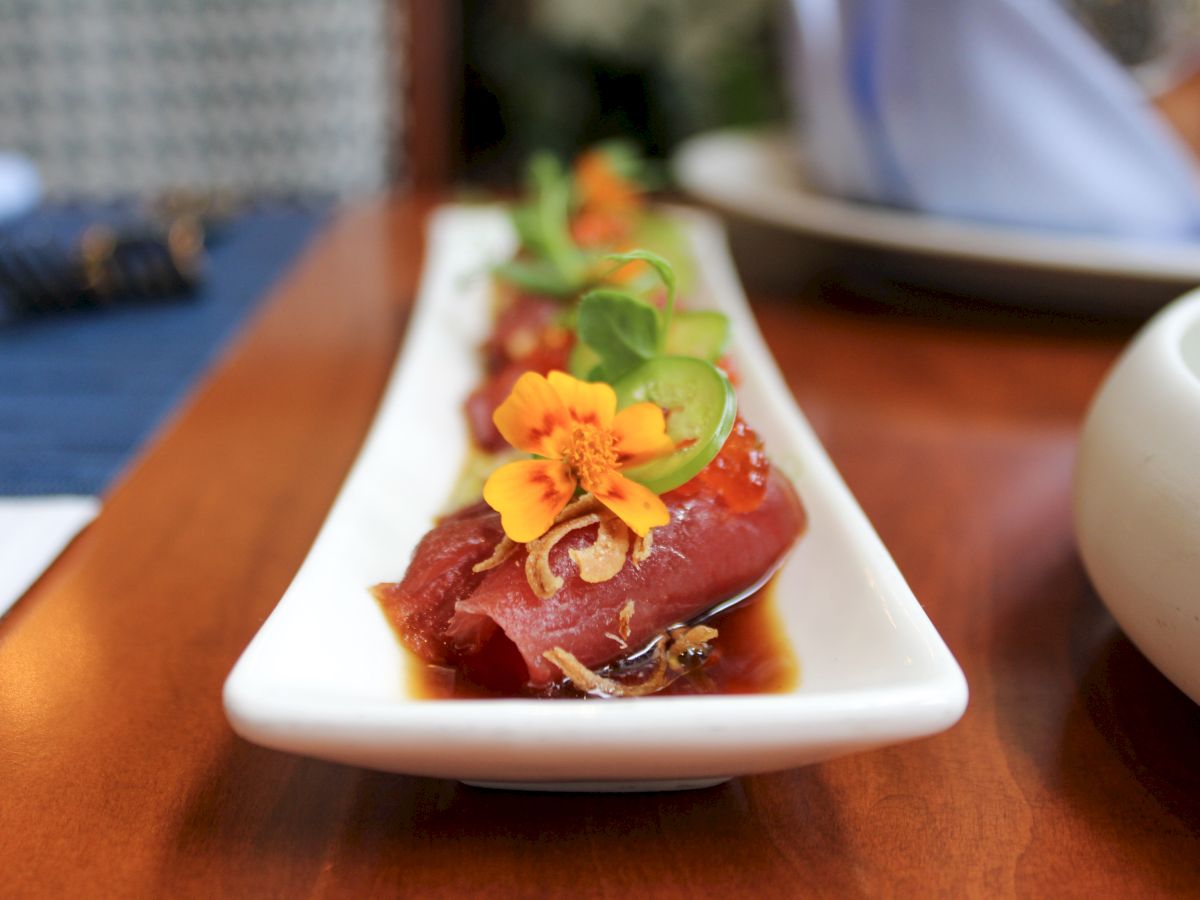 Sashimi pieces on a white rectangular plate, garnished with microgreens and edible flowers, placed on a wooden table.