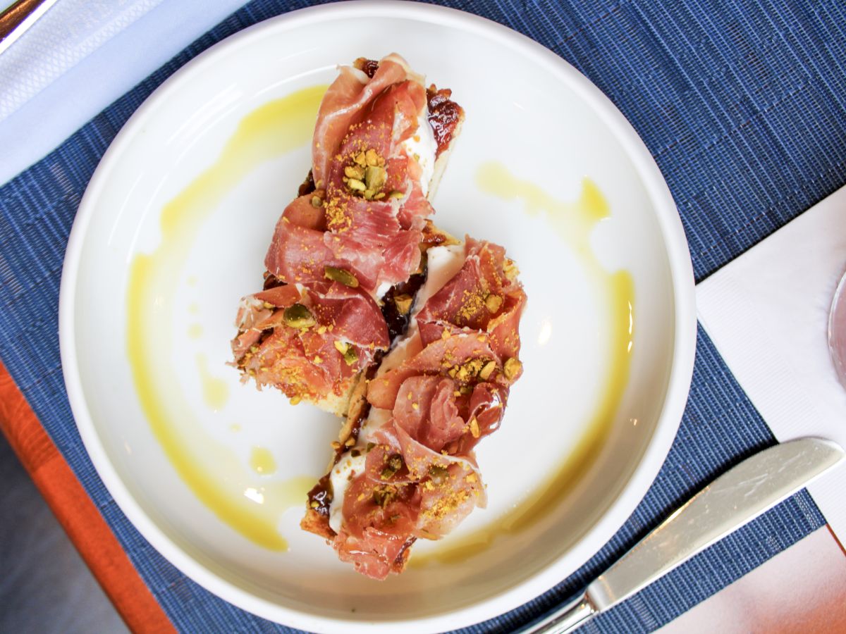 A white plate with two pieces of bruschetta topped with prosciutto and nuts, drizzled with olive oil, on a blue placemat with a knife and napkin.