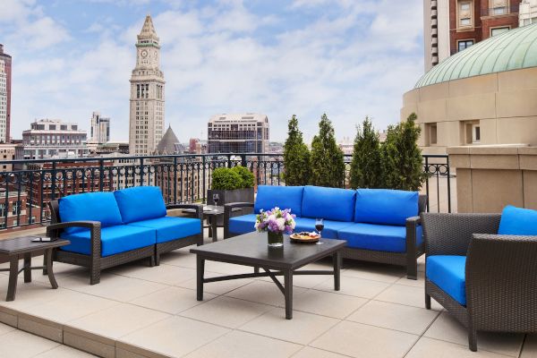 Rooftop patio with blue sofas, tables, and plants, overlooking a city.