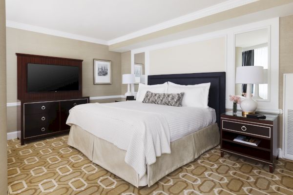 The image shows a neatly arranged bedroom with a large bed, two nightstands, a dresser with a TV, lamps, and a framed picture on a beige wall.