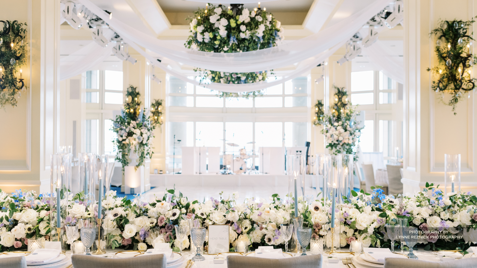 A beautifully decorated event space with floral arrangements, draped fabric, tall candles, and elegant table settings, likely for a wedding or reception.