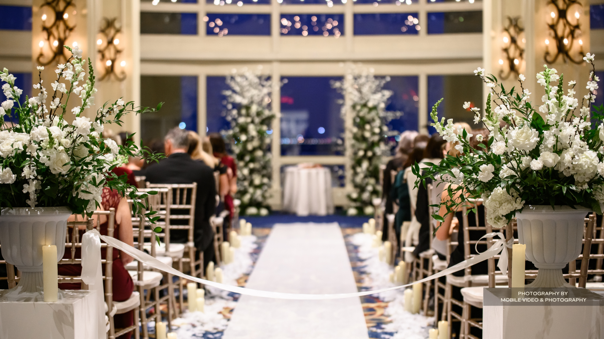 The image shows a beautifully decorated indoor wedding ceremony with white flowers, seated guests, and a floral aisle leading to the altar.