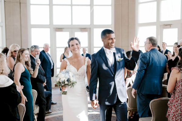 A smiling couple in wedding attire walks down an aisle, greeted by applauding guests in a bright room full of windows.