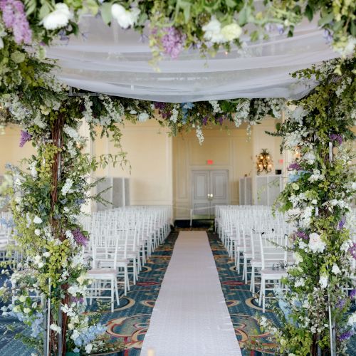 A floral archway leads to an aisle lined with white chairs in a beautifully decorated indoor wedding venue ending the sentence.