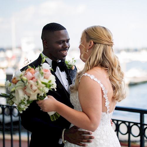 A couple in wedding attire smiles at each other near a waterfront, holding a bouquet.