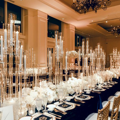 The image shows an elegantly set dining table with white flowers, tall candleholders, fine china, and gold chairs in a luxurious event space.