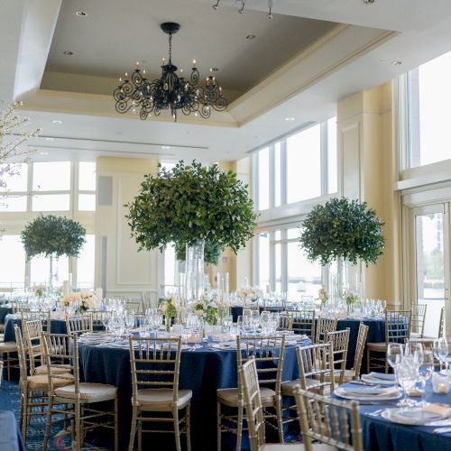 An elegant banquet hall set up for an event with round tables, gold chairs, floral arrangements, and large windows providing natural light.