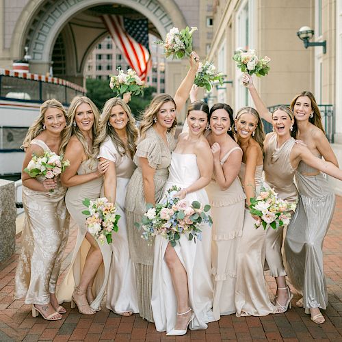 A group of women in formal dresses holding bouquets, smiling and posing joyfully outdoors with a large archway and flag in the background.