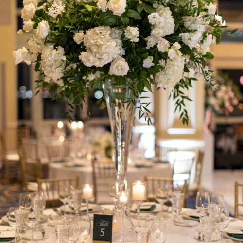 A elegantly set table with a tall floral centerpiece, white flowers, gold chairs, place settings, and a small table number 5 sign is pictured.