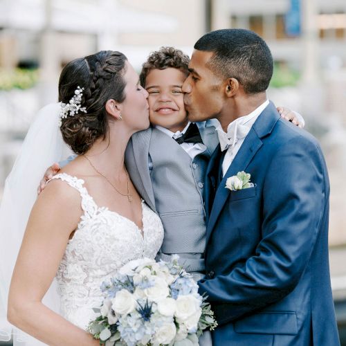 A bride and groom in wedding attire kiss a joyful child they are holding in between them, with the bride holding a bouquet.