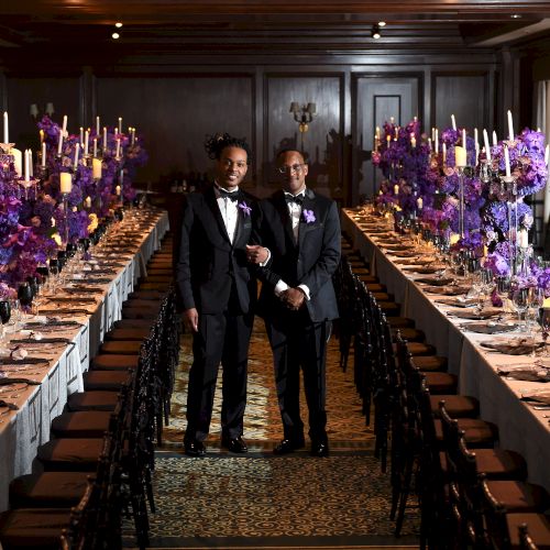 Two men wearing suits stand in an elegantly decorated dining hall with long tables, adorned with purple flowers and candles.