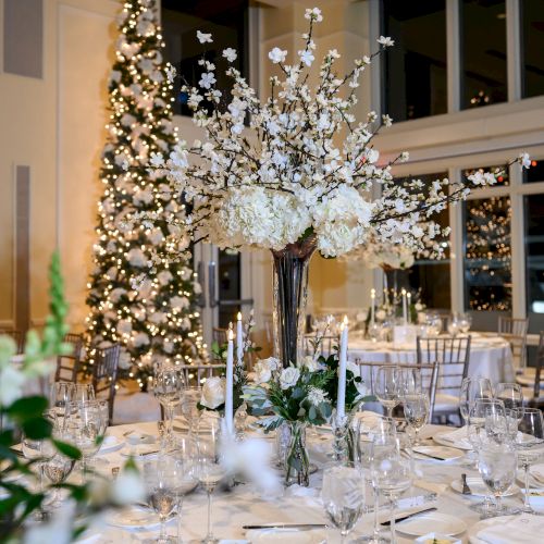 The image shows an elegantly decorated dining area with a large floral centerpiece, set tables, and a lit-up Christmas tree in the background.