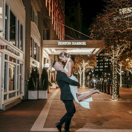 A couple is outside at night, with the man lifting the woman high in a joyful pose outside a well-lit building adorned with trees and lights.