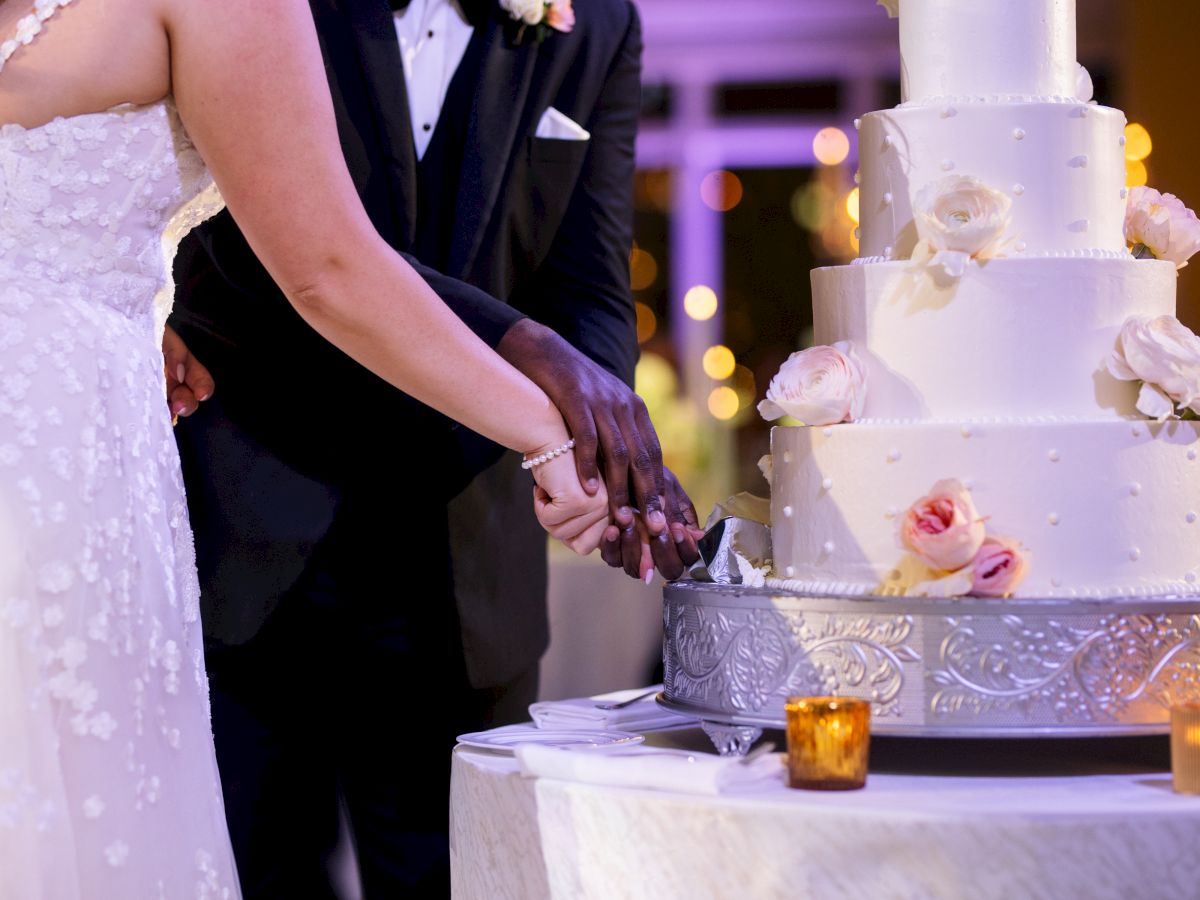 A couple is cutting a tiered wedding cake adorned with roses. They are in wedding attire, and small candles decorate the table.