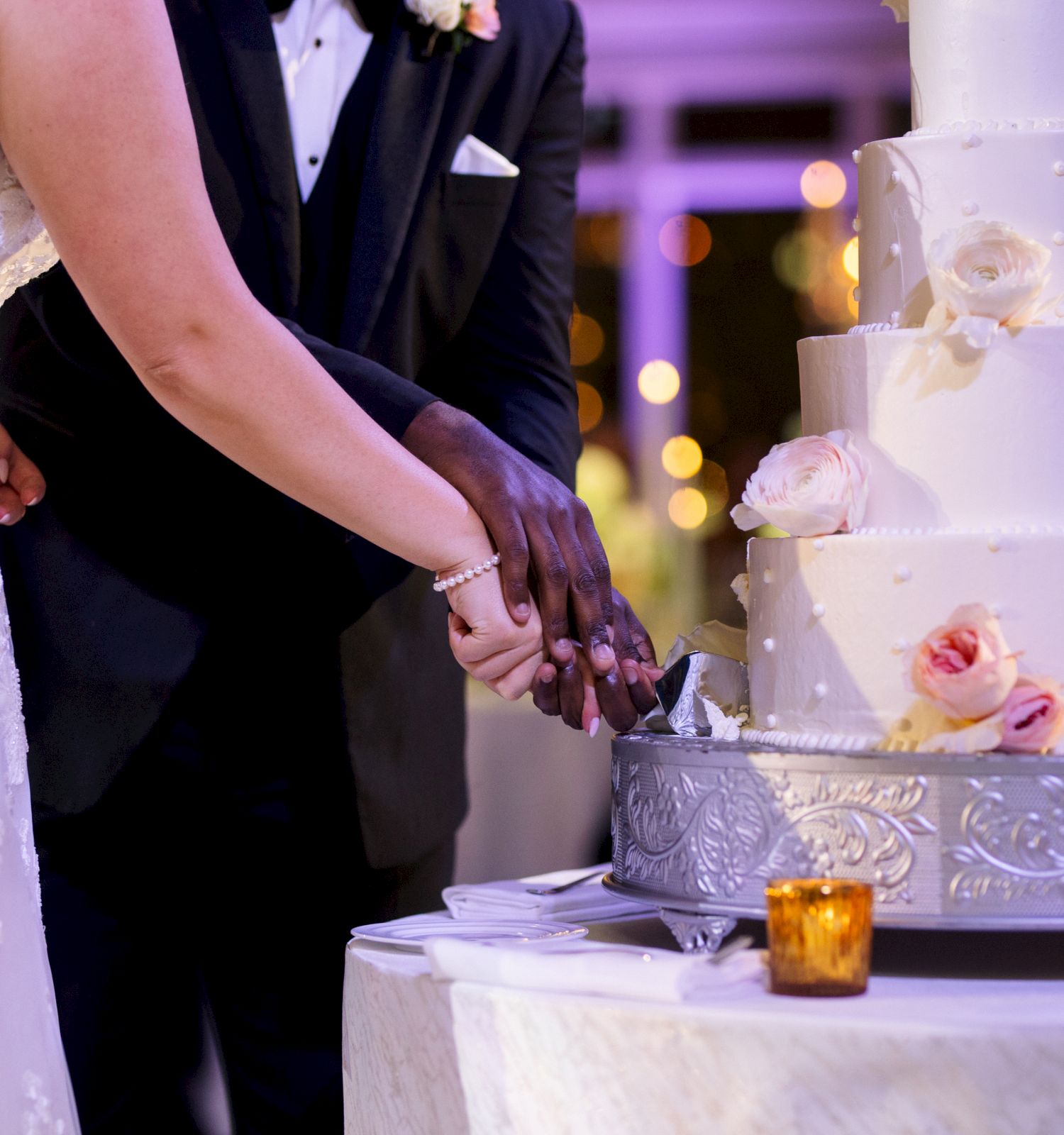 A couple is cutting a tiered wedding cake adorned with roses. They are in wedding attire, and small candles decorate the table.