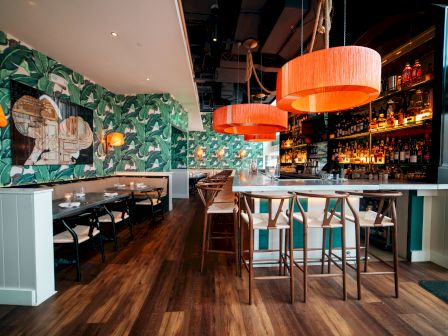 A modern restaurant with wooden floors, tropical wallpaper, and orange hanging lamps. The space includes a bar, high tables, and cozy booths ending.