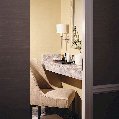 The image shows a makeup vanity with a cushioned chair, a countertop with various makeup products, a mirror, a wall lamp, and a small plant.