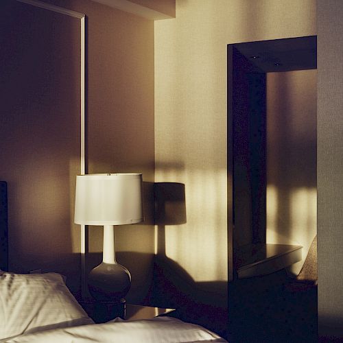 The image shows a bedroom with soft lighting. There is a bed with a white pillow and bedding, a bedside table, and a lamp next to a mirror reflecting light.