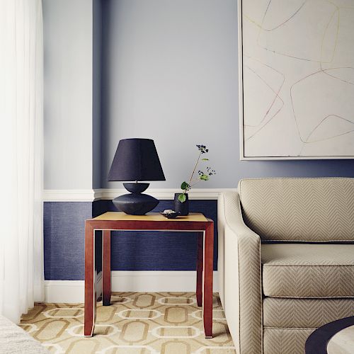 A living room corner with a beige sofa, a wooden side table with a black lamp and a small plant, and an abstract painting on the wall.