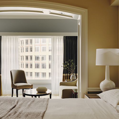 This image shows a cozy bedroom with a neatly made bed, a large window, two chairs, a round table, and a white lamp, creating a warm atmosphere.