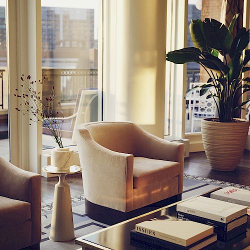 A modern living room with two armchairs, a coffee table with books, and large windows overlooking a cityscape is pictured here.