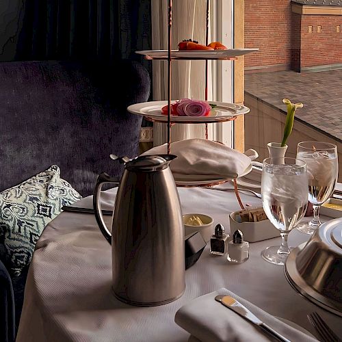 A table set for tea with a tiered tray of pastries, a metal pitcher, water glasses, and a vase with a single flower by the window.