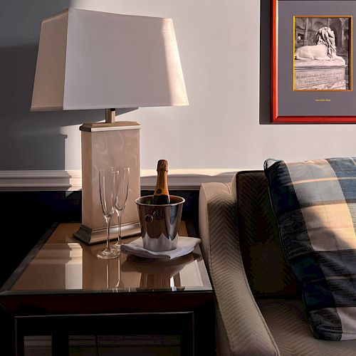 A cozy living room corner with a lamp on a side table, a sofa, wall art, and a champagne bottle in an ice bucket.