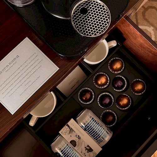 A coffee station with a Nespresso machine, coffee pods, two cups, bottled water, and a card on a wooden surface.