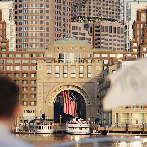 A person looks toward a grand archway with an American flag, surrounded by tall buildings and a waterfront area with boats docked.