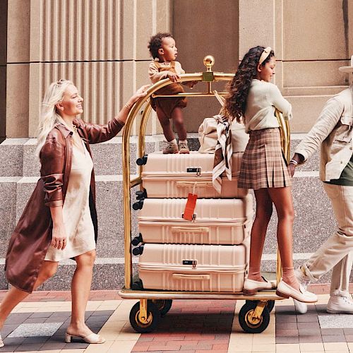 A stylish group, including a child, pulling a luggage cart stacked with suitcases on a city sidewalk with columns in the background.