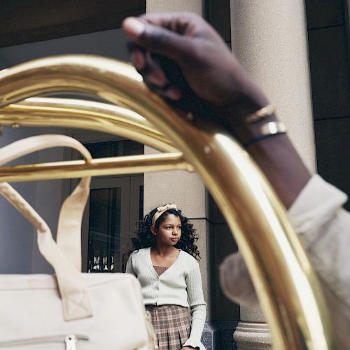 A person with a bag stands in the background near a column, while another person holds a hotel luggage cart in the foreground, framing the scene.