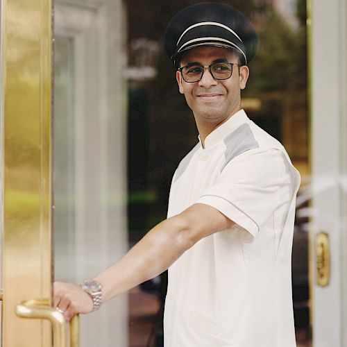 A doorman in a white uniform and a cap is holding a door open while smiling at the camera.