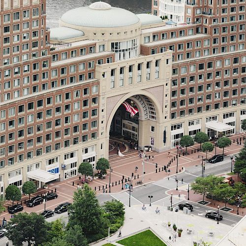 Aerial view of a large building with a prominent archway entrance, surrounded by greenery and intersecting roads. Buildings and waterfront in the background.