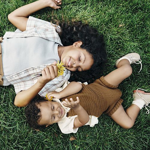 Two children are lying on the grass, smiling, and holding yellow flowers. One child has a plaid shirt and the other is dressed in brown.