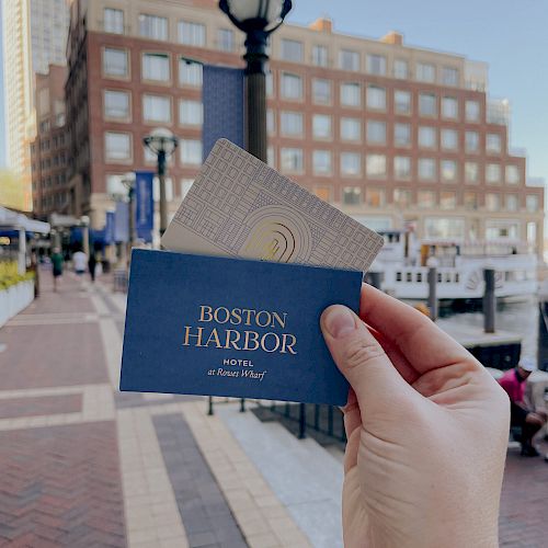 A hand holds a "Boston Harbor" card in front of a waterfront building with a lamppost, walkway, and people in the background.