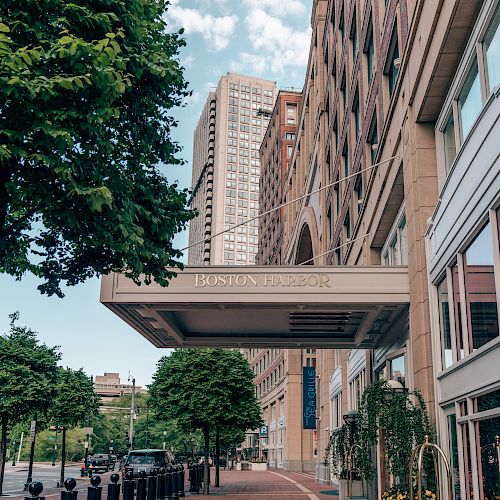 The image shows a city sidewalk with trees, a large overhang structure, and multi-story buildings in the background under a blue sky with clouds.