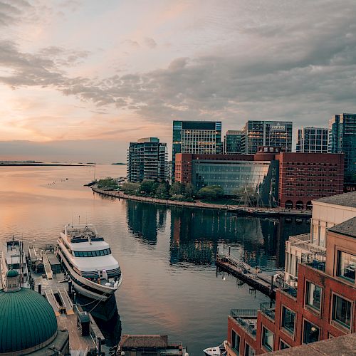 A waterfront view with modern buildings, a docked boat, and calm water under a cloudy sky at sunset.