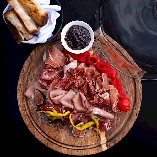 A charcuterie board with various cured meats, figs, and peppers next to a bowl of sauce and a basket of toasted bread is served.