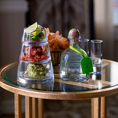 The image shows a table with chips, salsa, guacamole, a bottle of tequila, lime garnishes, and two shot glasses arranged on a clear glass surface.