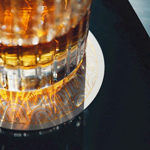 A close-up of a glass filled with a golden-brown liquid, likely whiskey, sitting on a round white coaster on a dark surface.