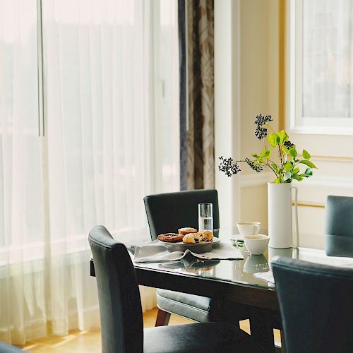 A dining area with a table set for breakfast, including a plate of food and a glass of water, surrounded by black chairs, and a window with curtains.